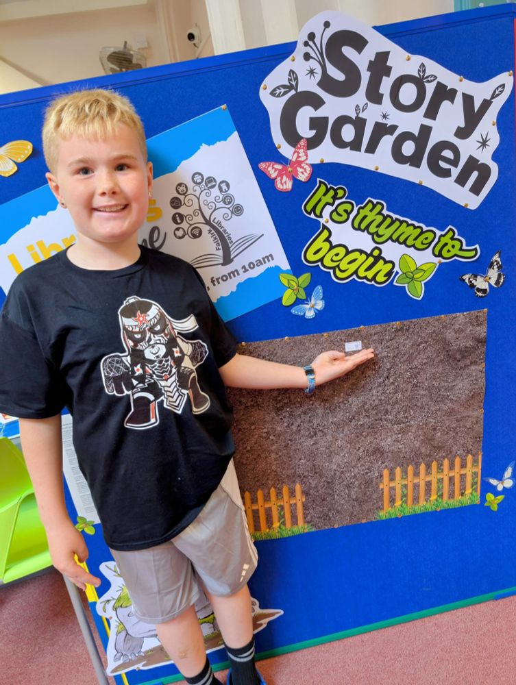A primary age boy with short blonde hair is smiling in front of a board display that has the caption "Story Garden".