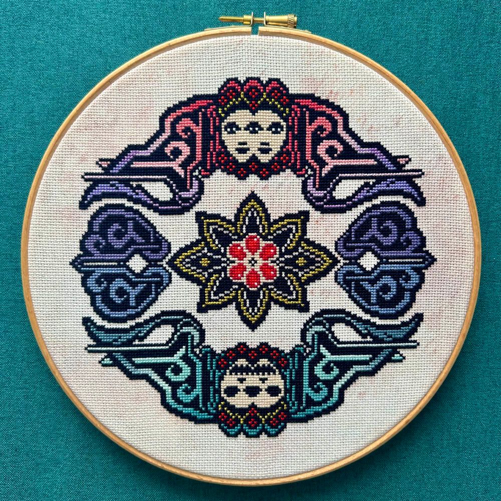 Pattern designed and stitched by Screaming Heart Design.

A geometric design mirrored vertically. Top half features a woman’s face with warm purples and pinks. Bottom half features a skull with cool teals and blues. A pomegranate with six seeds sits in the middle.