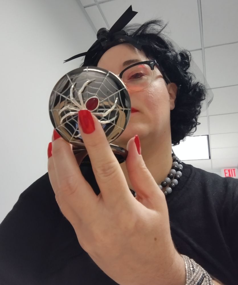 a person wearing all black. Their nails are very red and they are checking their makeup in a compact with a spider on it. 