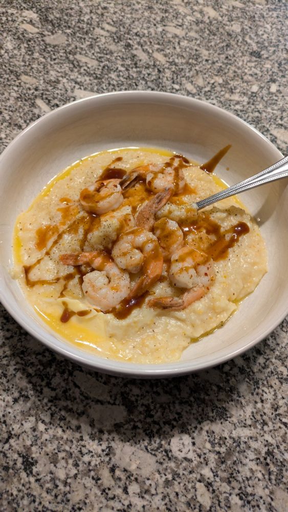 Bowl of grits topped with sauteed shrimp, tonkatsu, and hot sauce on a granite countertop
