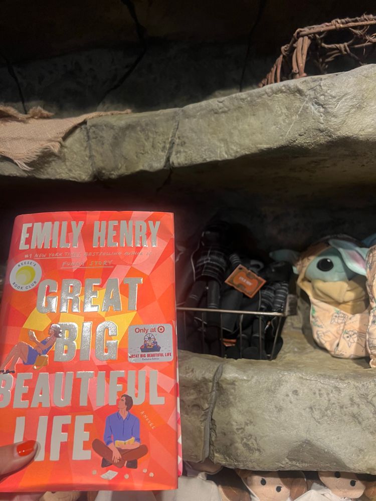 Emily Henry’s book “Great Big Beautiful Life,” whose cover characters resemble modern AU Ben and Rey from Star Wars, in the foreground with a Kylo Ren plushie in the background in downtown Disney at the Star Wars Trading Post store  