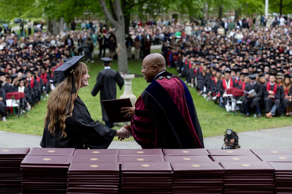 With the Class of 2025 seated on the Historic Quad in the background we see Bates President Garry W. Jenkins, on right, hand over a diploma and shake hands with a smiling Bates student, on left. 