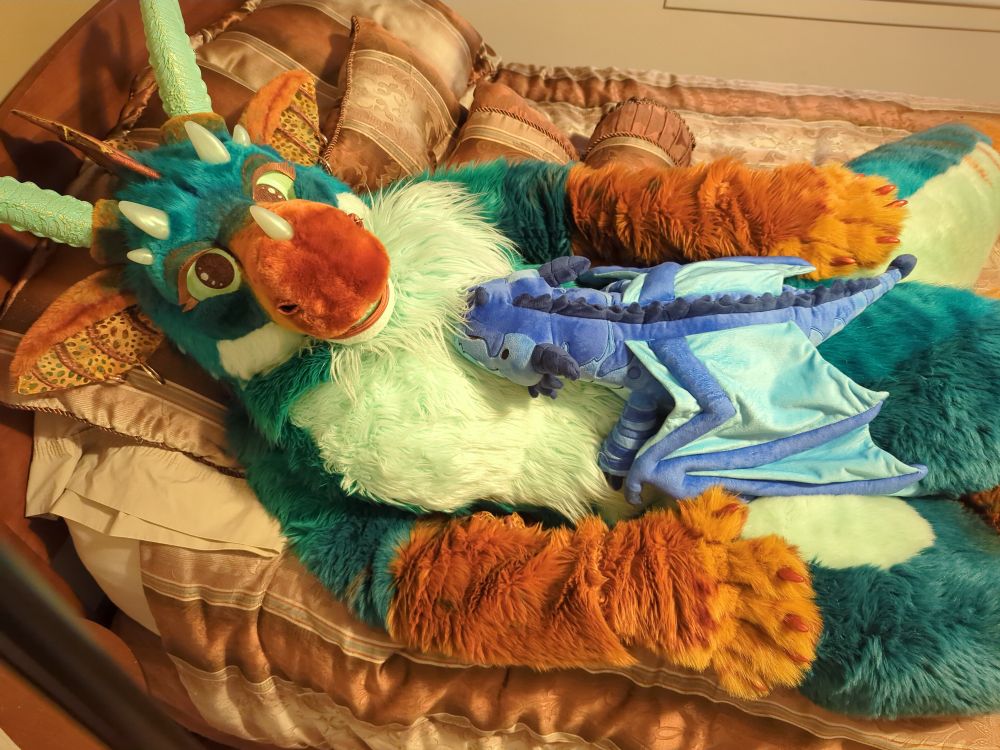 Avala fursuiter lying on a bed with a blue dragon plush lying on her stomach.