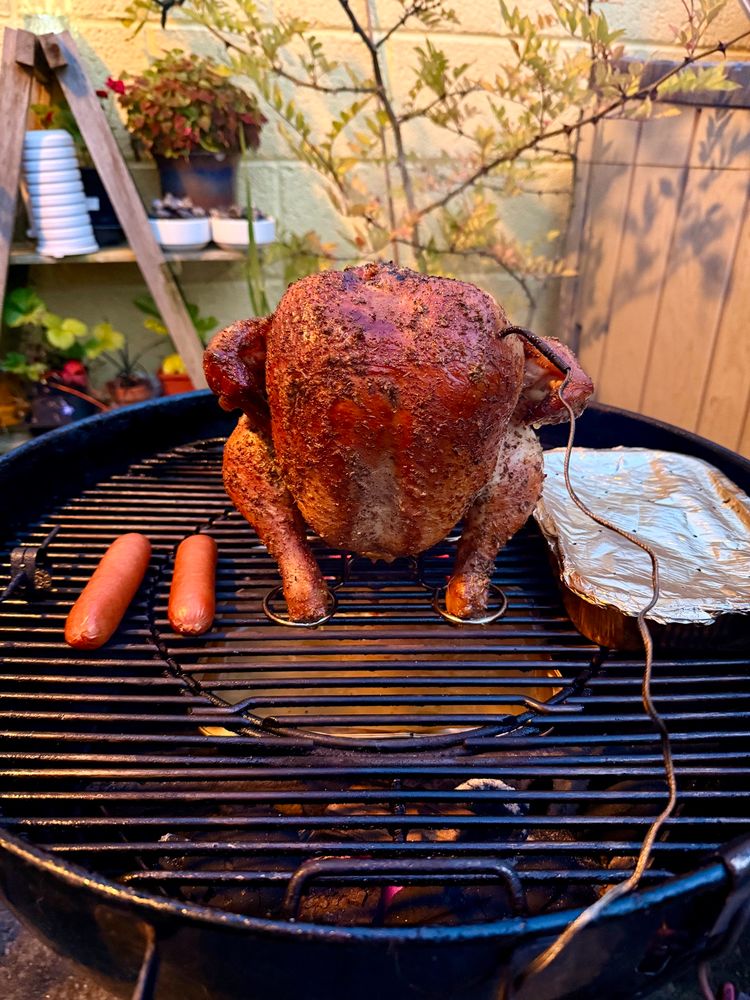Chicken standing in a beercan holder on a Weber kettle, flanked by a couple of sausages & foil tray of baby potatoes; all bathed in a smoked glow of hickory smoke