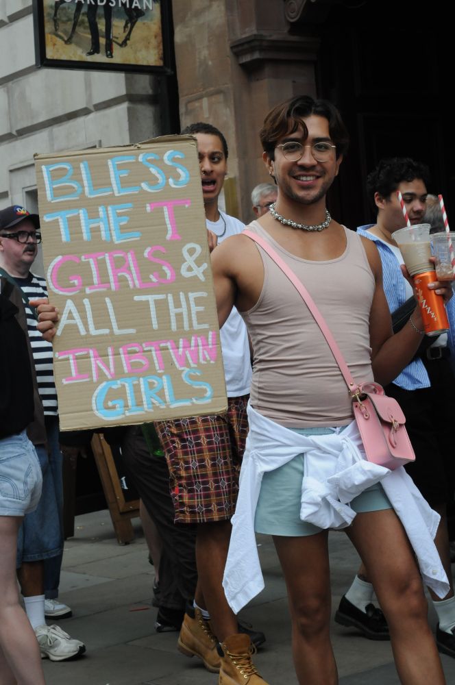 Person wearing beige vest and green hot pants holds sign that says bless the t girls and all the in between girls