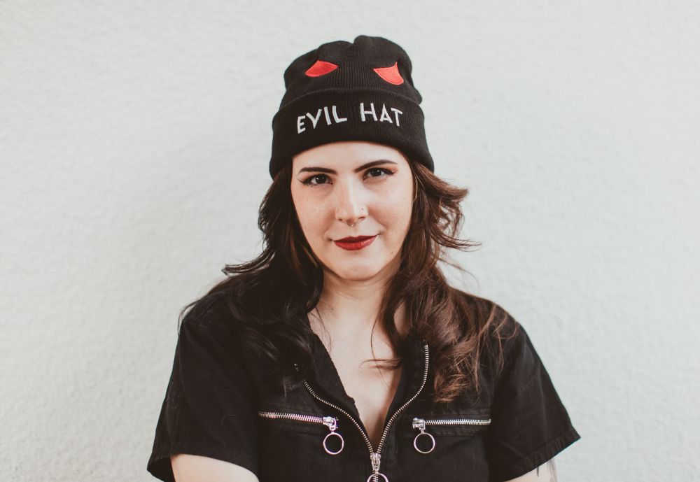 A woman with long brown hair wearing a black jumpsuit with big shiny zippers. Atop her head is a black beanie embroidered with red "evil" eyes and the words "Evil Hat" across the bottom brow. 