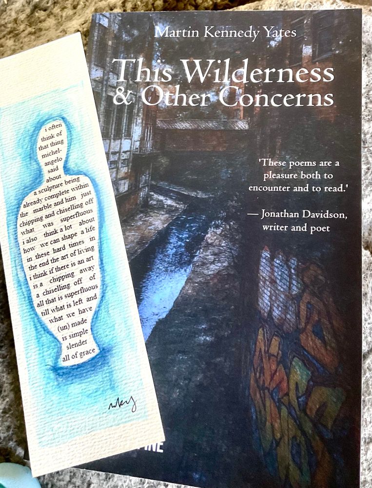 A photo of a poetry book and a bookmark. The book is dark blue in colour and has a painting of a wet city street with partial buildings visible an a wall on the right with graffiti. Text as follows

Martin Kennedy Yates
This Wilderness 8c Other Concerns
'These poems are a pleasure both to encounter and to read.'
- Jonathan Davidson, writer and poet