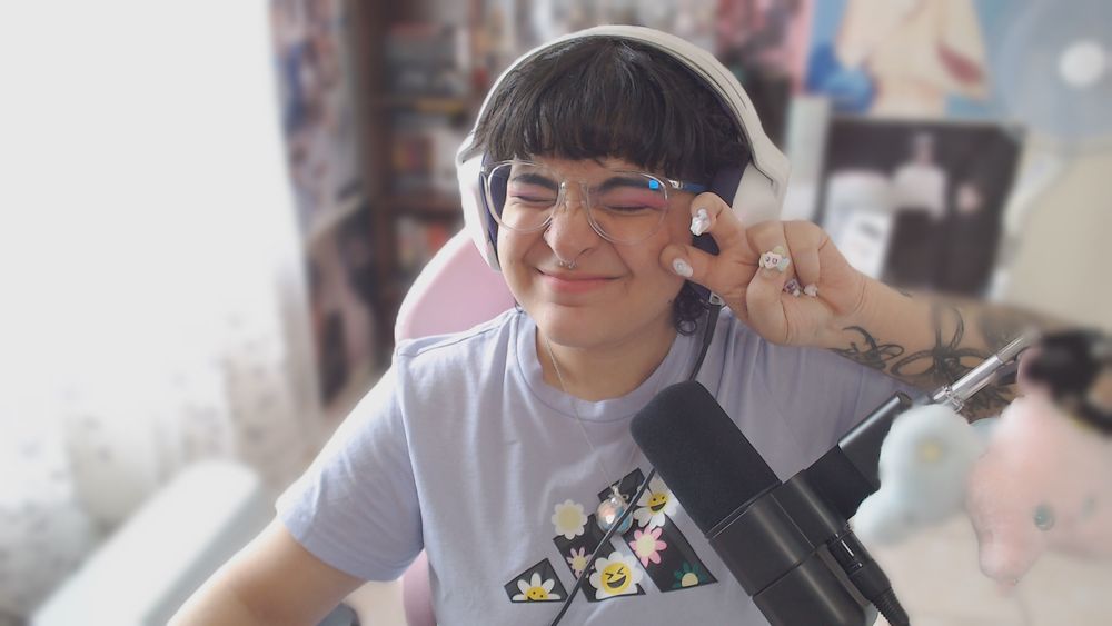 menji holding up a peace sign and smiling with their eyes closed. they’re a lightskinned latine with dark black short hair, they wear glasses and a lilac/blue t-shirt.