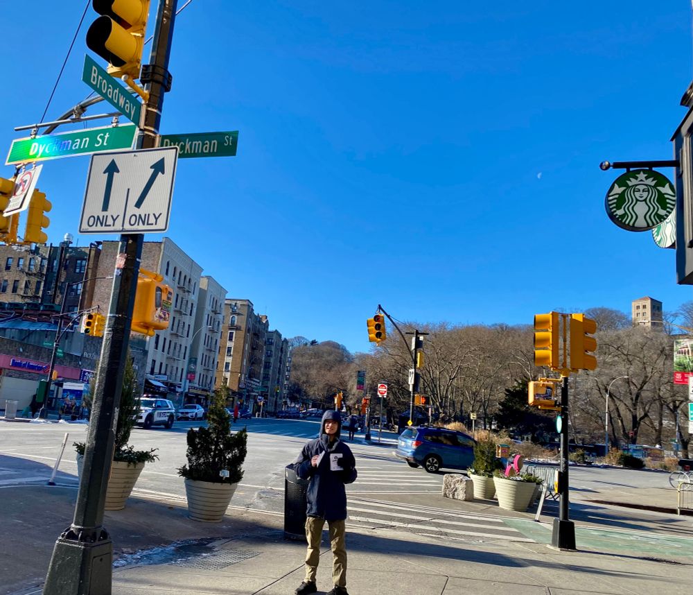 A young man in a warm coat stands on an urban street corner next to a Starbucks sign. Street signs indicate that  it is the corner of Broadway and Dyckman St. The young man is holding out flyers in support of striking Starbucks workers.