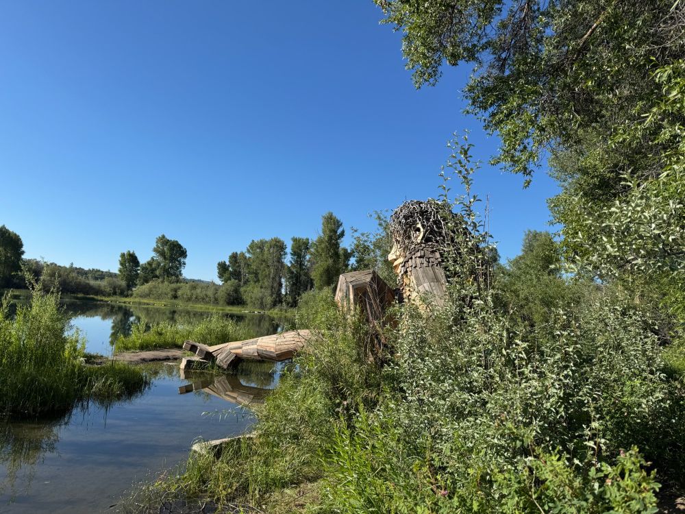 Mama Mimi - a large wooden troll built by the Danish artist, Thomas Dambo. She sits on the shore of a clear lake with one leg extended over the water. The sky is bright blue, the trees and foliage are spring green, and the lake is reflecting it all.