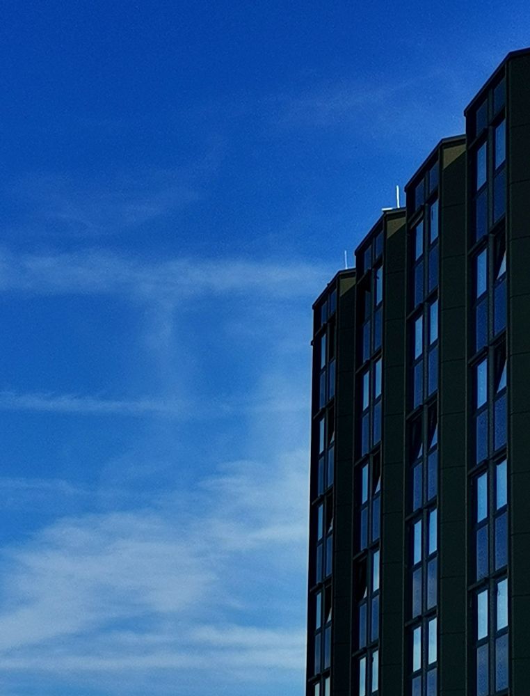 a simple photography. It's a bright blue sky with some soft, fog like clouds. On the left side of the picture is a big building with 4 rows next to each other's. Each row has two windows next to each other. The floors are switching between windows and wall. The building over all is a new, modern one. It's most likely blue, because of the reflection from the sky