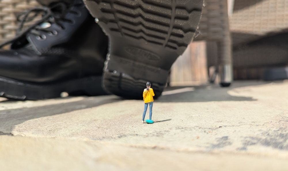 A tiny figurine of a black-haired woman in a yellow jacket and jeans is dwarfed by my boot sole.