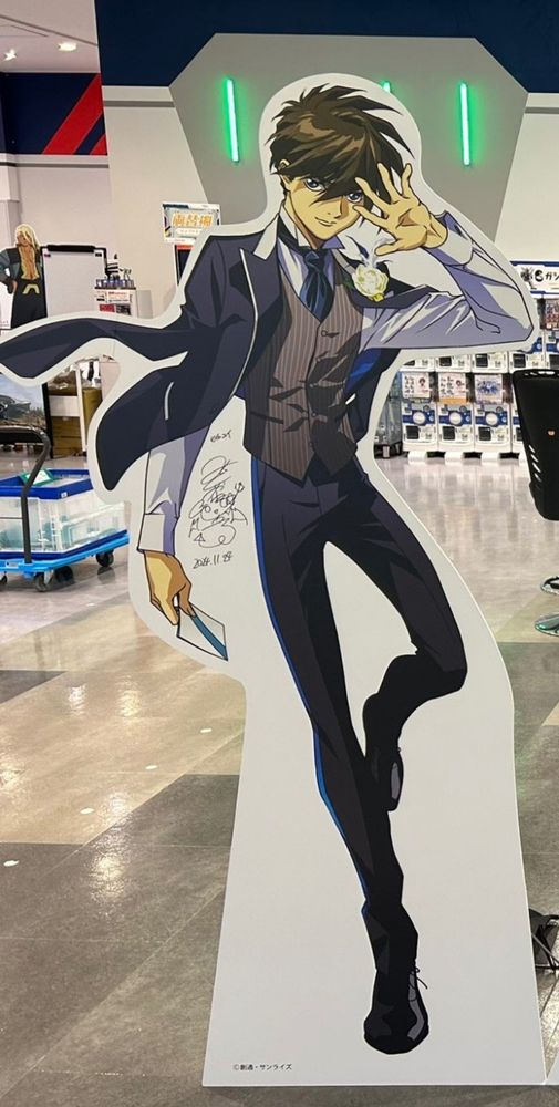 A cardboard cut out of Heero Yui from Gundam Wing wearing a fitted suit with the jacket hanging on his shoulders, and striking a handsome moving pose.