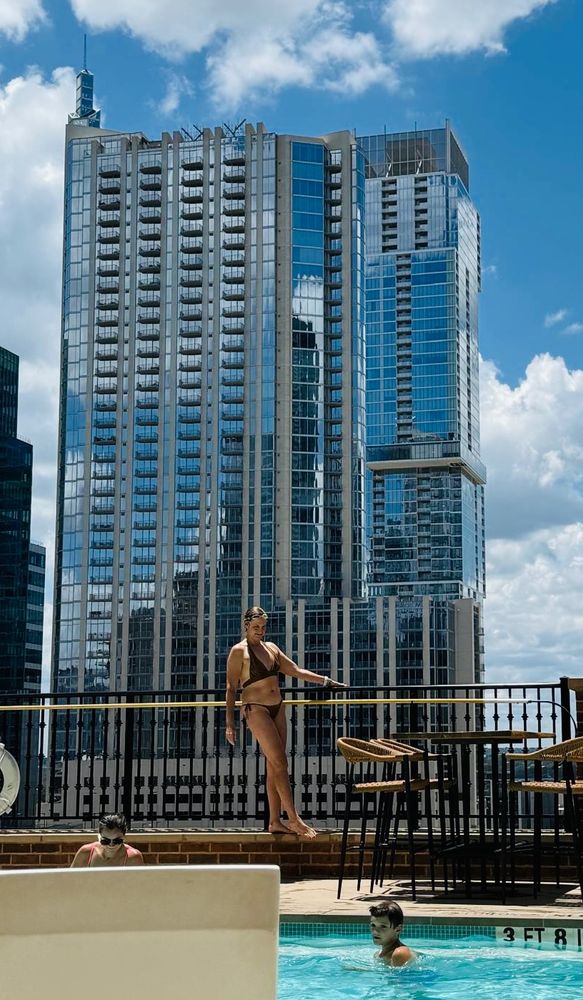 A pale woman in a black bikini poses with crossed legs against a railing with a large, mirrored skyscraper in the background
