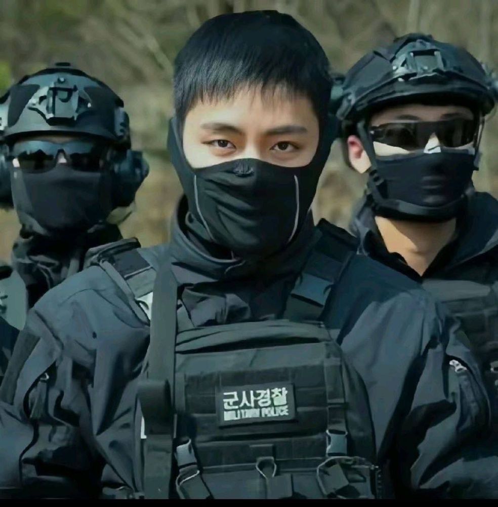 Três homens, sendo dois atrás e um na frente.
Todos usam o uniforme preto da Polícia Militar da Coréia do Sul. 
Os dois de trás estão de capacete, óculos escuros e máscara no rosto. 
O da frente é Kim Taehyung, o V do BTS, está usando apenas a máscara no rosto. É asiático, com olhos castanho escuros e cabelos pretos.