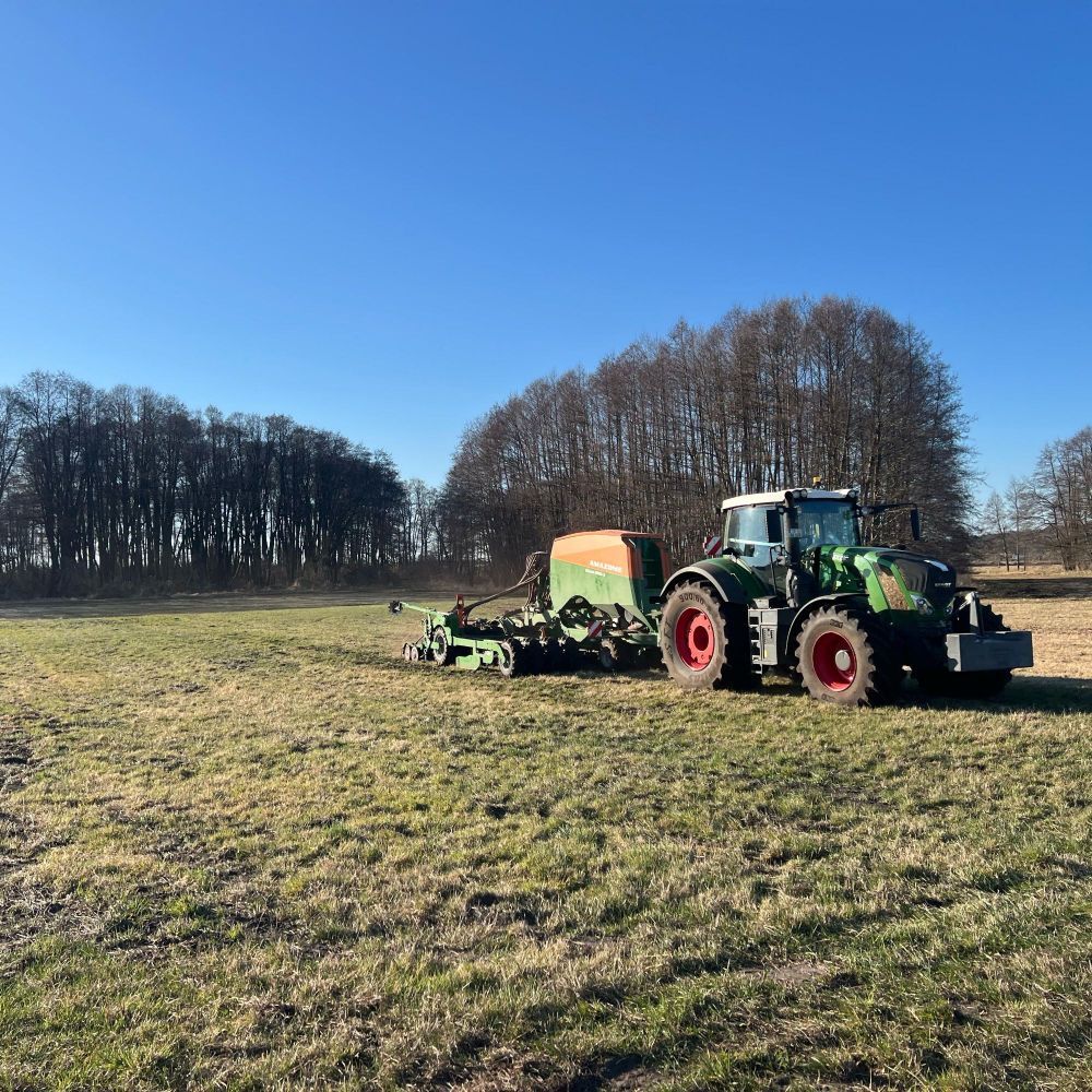 Ein Fendt-Traktor mit einer Drille von Amazone auf dem Grünland. Im Hintergrund sind Pappelstreifen zu sehen, die Struktur in die Landschaft bringen.