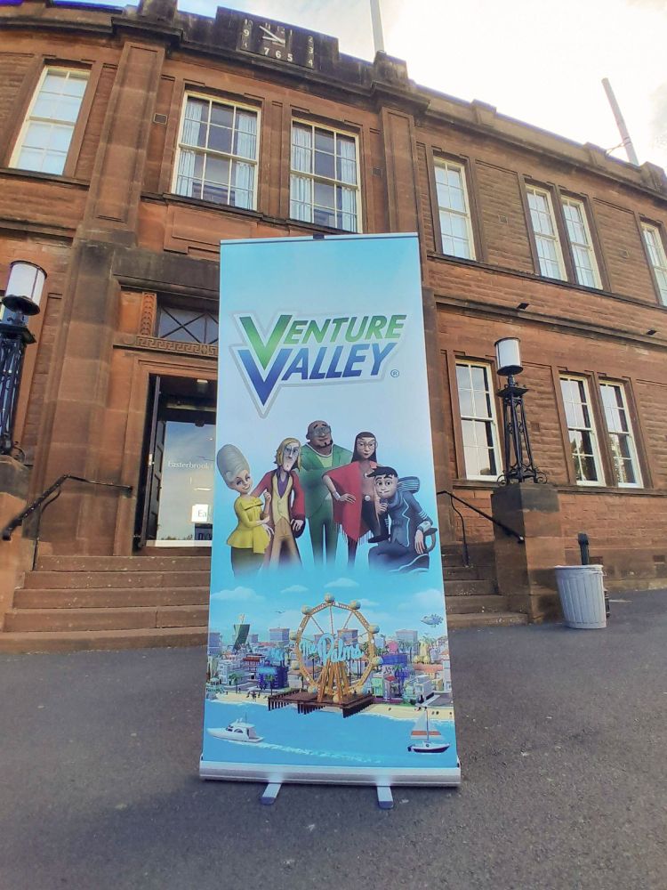 A large Venture Valley Poster on display in front of a grand red brick building.