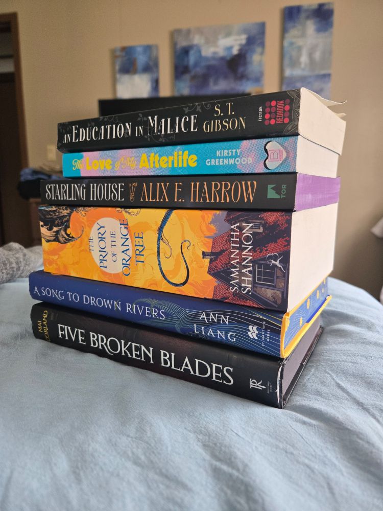 A stack of 6 books. The books are: An Education in Malice, The Love of My Afterlife, Starling House, The Priory of the Orange Tree, A Song to Drown Rivers, and Five Broken Blades.