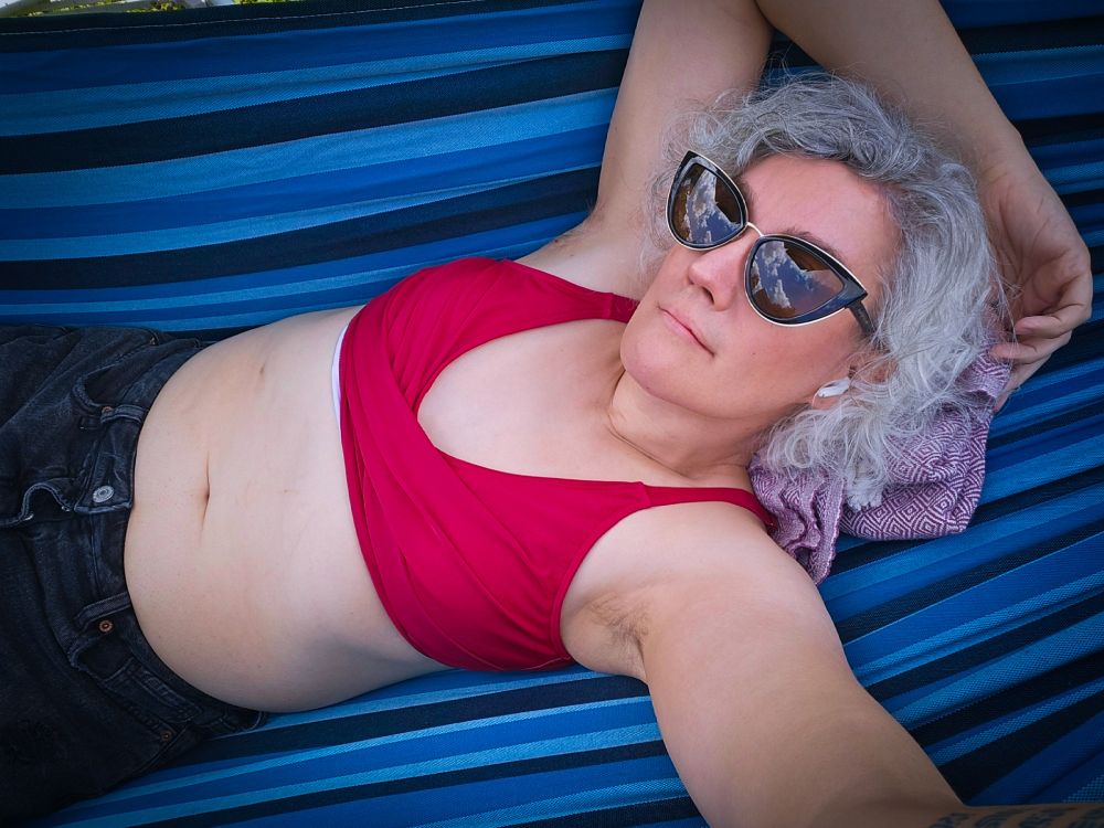 Alice, a white woman with curly gray hair and cats eye sunglasses, lays in a hammock wearing a red bikini top and black jean shorts