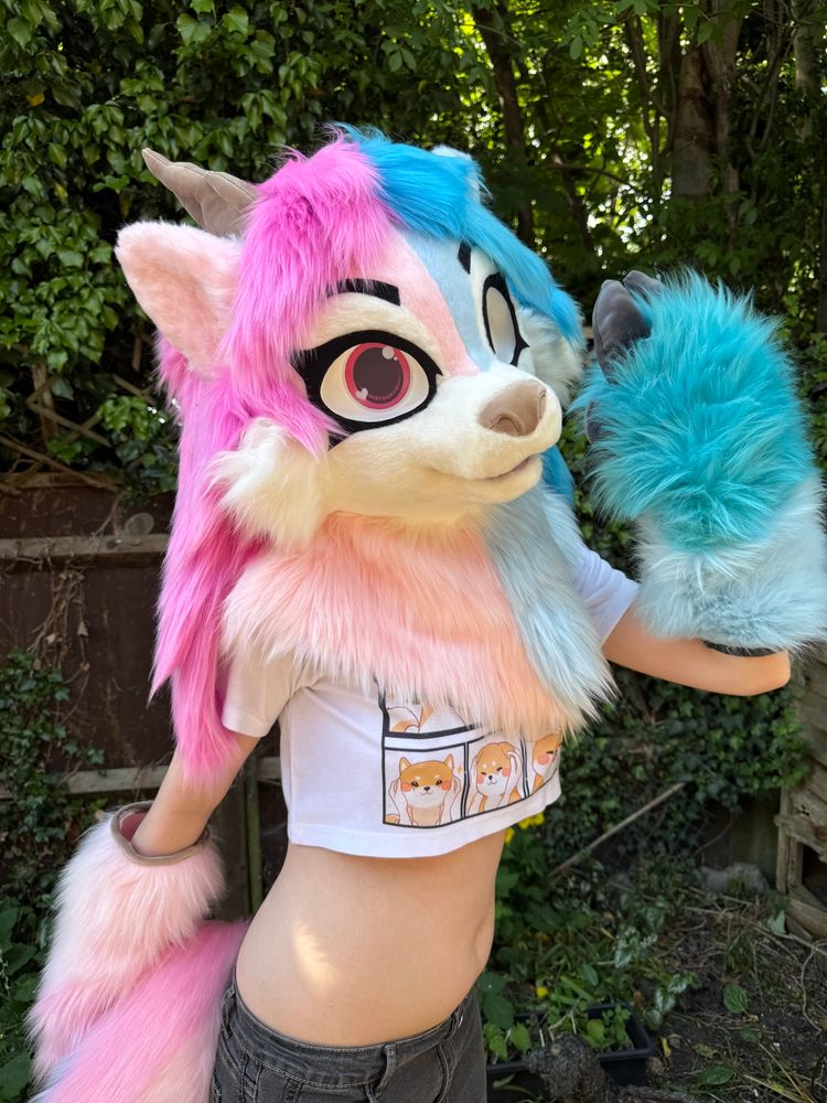 Blue and pink deer fursuit on display. The suit has long, feminine hair and blue and pink eyes. She has matching blue and pink hoof paws and brown and grey horns. She is posed with one hand raised. 