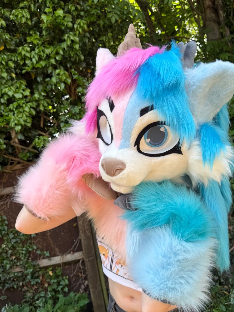 Blue and pink deer fursuit on display. The suit has long, feminine hair and blue and pink eyes. She has matching blue and pink hoof paws and brown and grey horns. She is posed with both hands raised to her chin.