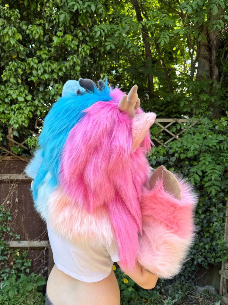 Blue and pink deer fursuit on display. The suit has long, feminine hair and blue and pink eyes. She has matching blue and pink hoof paws and brown and grey horns. She is posed with her back turned 