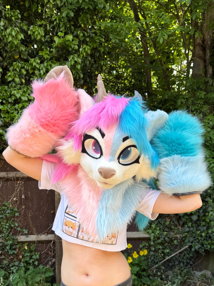 Blue and pink deer fursuit on display. The suit has long, feminine hair and blue and pink eyes. She has matching blue and pink hoof paws and brown and grey horns. She is posed with hands above her head 