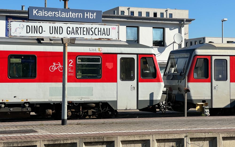 Zwei VT 628 im Sylt Shuttle Plus Design am Bahnsteig. Am Bahnsteig das Bahnhofsschild „Kaiserslautern Hbf“, darunter ein weißes Zusatzschild „DINO- UND GARTENSCHAU“