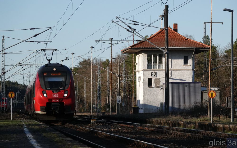 Bei untergehender Sonne: Herannahender Talent 2 Zug neben einem von der tiefstehenden Sonne angeleuchteten, weißen Stellwerksgebäude mit roten Ziegeln, umgeben von Wald.