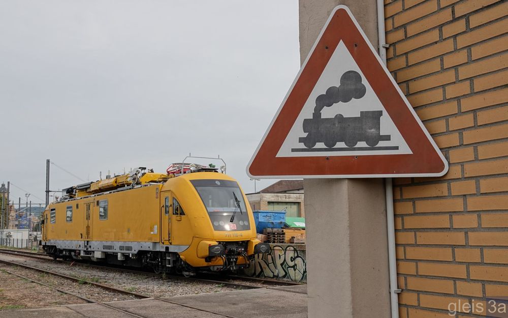 Im Vordergrund ein Verkehrszeichen "Bahnübergang" in älterer Ausführung mit Dampflok-Piktogramm. Im Hintergrund ein Instandhaltungsfahrzeug DB-Baureihe 711.1.