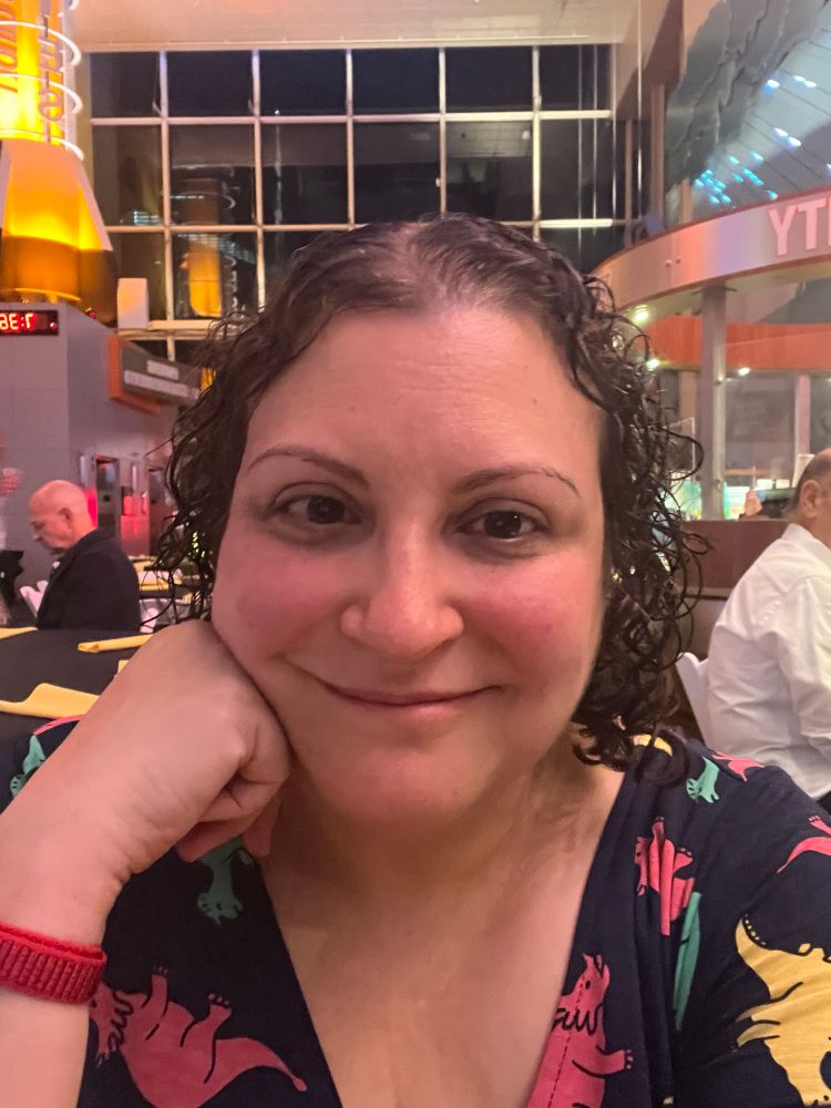 A middle-aged woman with brown curly hair, wearing a navy dress with colorful dinosaurs on it.