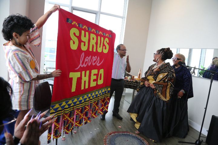 Milo and lwrds hold up a pink and neon yellow banner that says syrus loves Theo while syrus and Theo look on