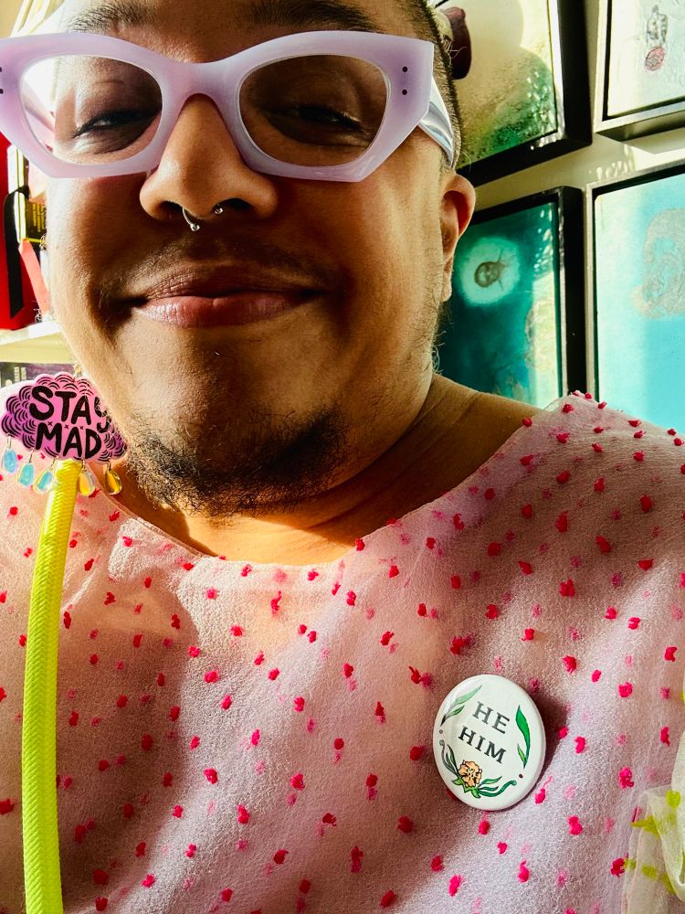Syrus wears a hello darling dress and a he/him button and lavender glasses and smiles at the camera 