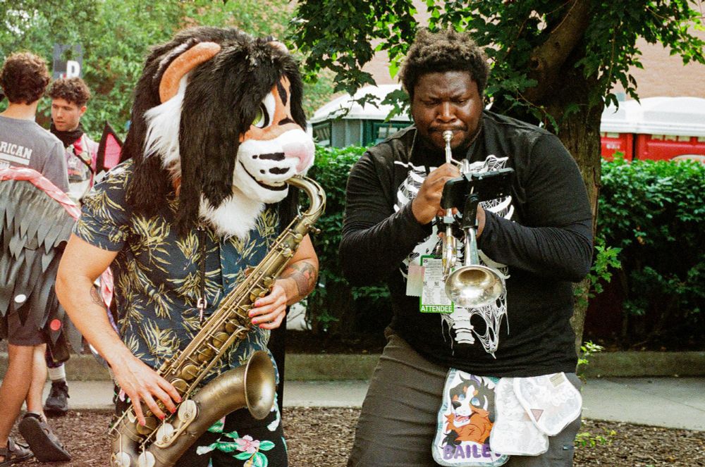 A person in a tiger fursuit head playing the sax, joined by another person playing the trumpet
