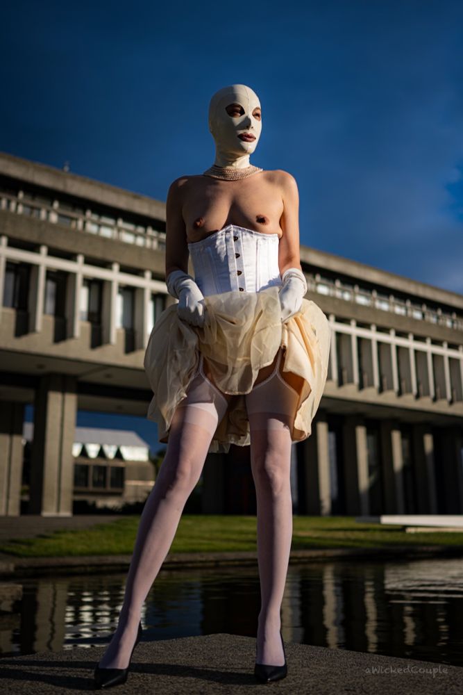 The rubberdoll in white stands, legs apart, lifting her skirt to reveal her white stocking tops across her thighs, in front of some brutalist architecture and a reflecting pool.  AWickedCouple Photography. 