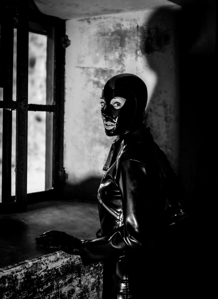 The black and white photo shows sharp contrasts of light and stark shadows as the rubberdoll dressed in black latex stands at a window, but with eyes turned back towards the camera, almost as if she has been caught by the light performing an illicit act. aWickedCouple Photography. 