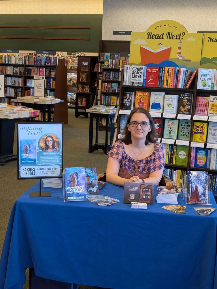 A picture of Millie Abecassis, a 35-yo white woman with shoulder-length brown hair, brown eyes, glasses, and wearing a pink and purple top, sitting at a table in a bookstore and smiling to the camera. The table is covered with books, bookmarks, and other items. Behind are shelves of books.
