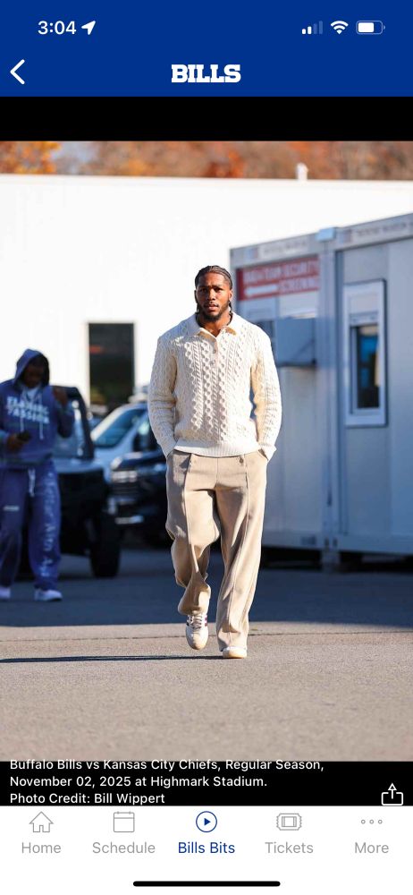 A Buffalo Bills player wearing a really cool off-white cable-knit sweater with a polo collar, nicely-structured baggy camel-colored felted pants with exaggerated seaming down the front of the legs, and white leather Adidas with brown stripes.
