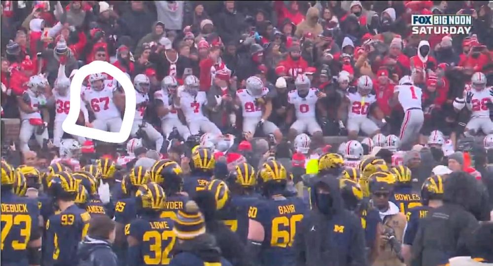 A picture of Ohio State players celebrating their victory in the stands with the fans while Michigan players guarded the M at the center of their field for a flag planting that never happened. The Ohio State player wearing jersey #67 in the stands is circled. 