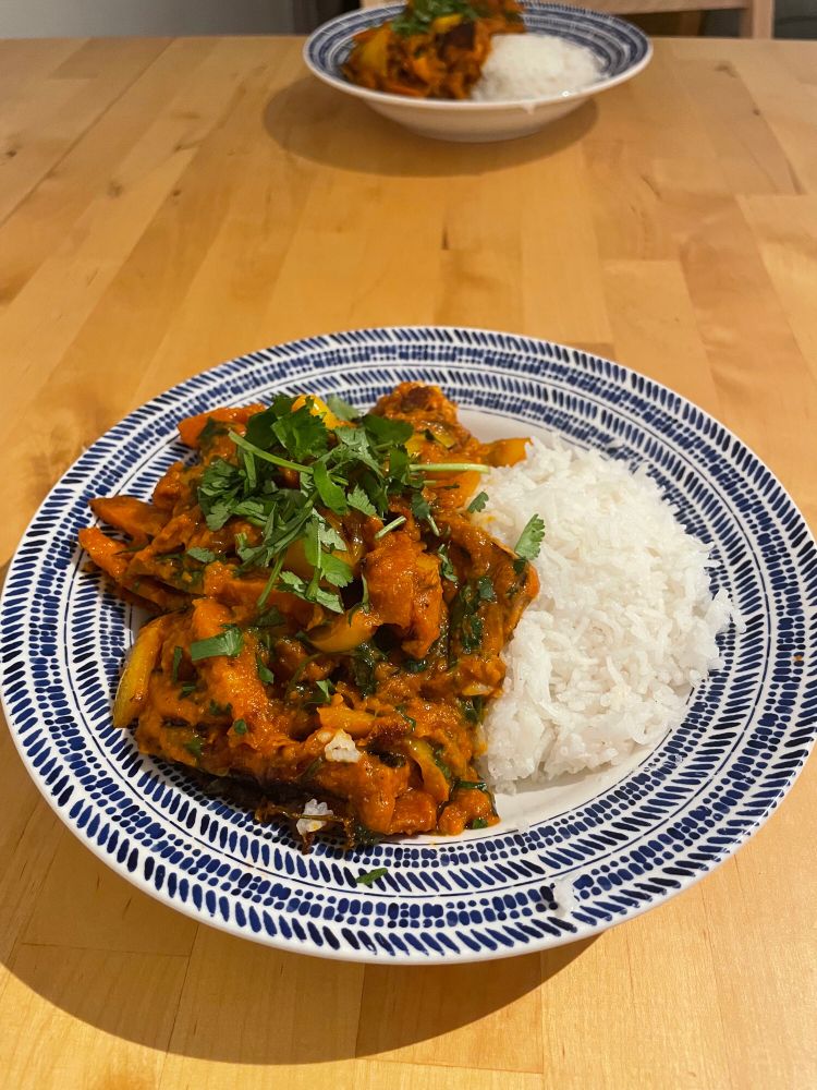 Two plates of food and a wooden table. A red-orange tofu and vegetable curry with white rice topped with green coriander leaves