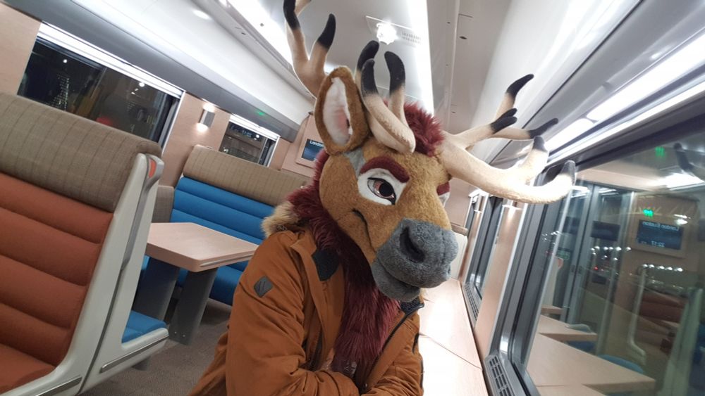 A selfie of a brown and red caribou fursuiter partialling in a fancy looking train dining car wearing a brown jacket.
