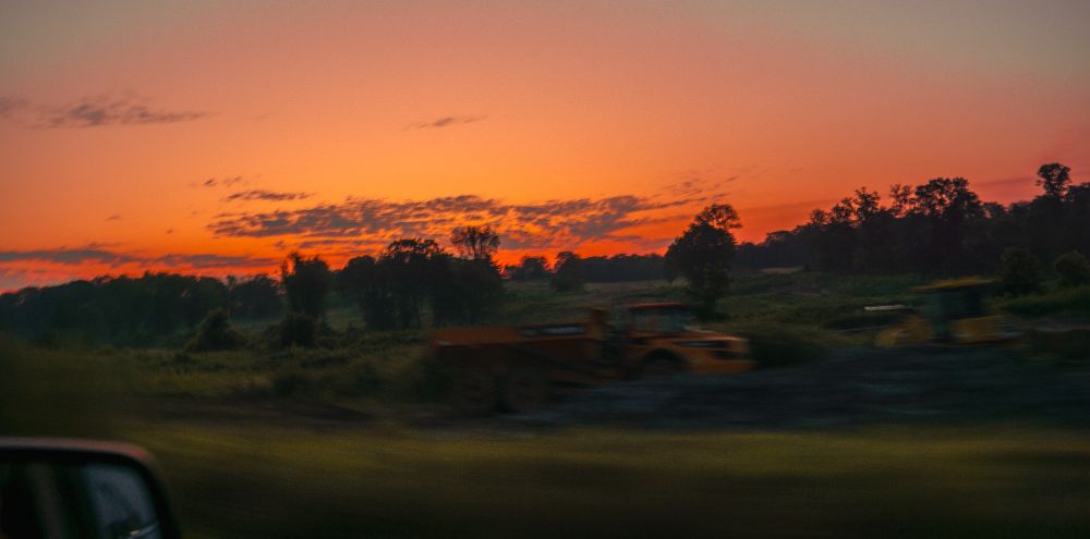 Before the sun breaks, dense orange hues stretch across the sky—concentrated at the horizon, diffusing upward into pale gradients. The orange is not decorative; it signals ignition. Below, silhouetted construction trucks and machinery hold scale and function, not blur. Greenery lines the edge of the site, unlit and unsoftened. The orange operates as temporal marker—before emergence, not after.