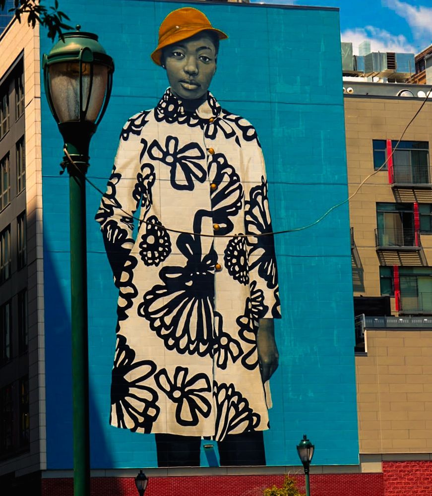 A mural of a woman in a yellow hat and black-and-white floral coat painted on a bright blue wall in downtown Philadelphia,  Buildings and a streetlamp are visible nearby.

