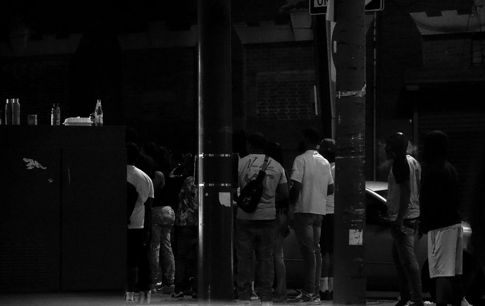 Nighttime city sidewalk near a utility box. Several people stand close together, with bottles and other items placed on the box. A pole runs through the center of the frame. Behind them is a brick wall and blurry signage. The lighting is dim, and a parked car is partially visible.
