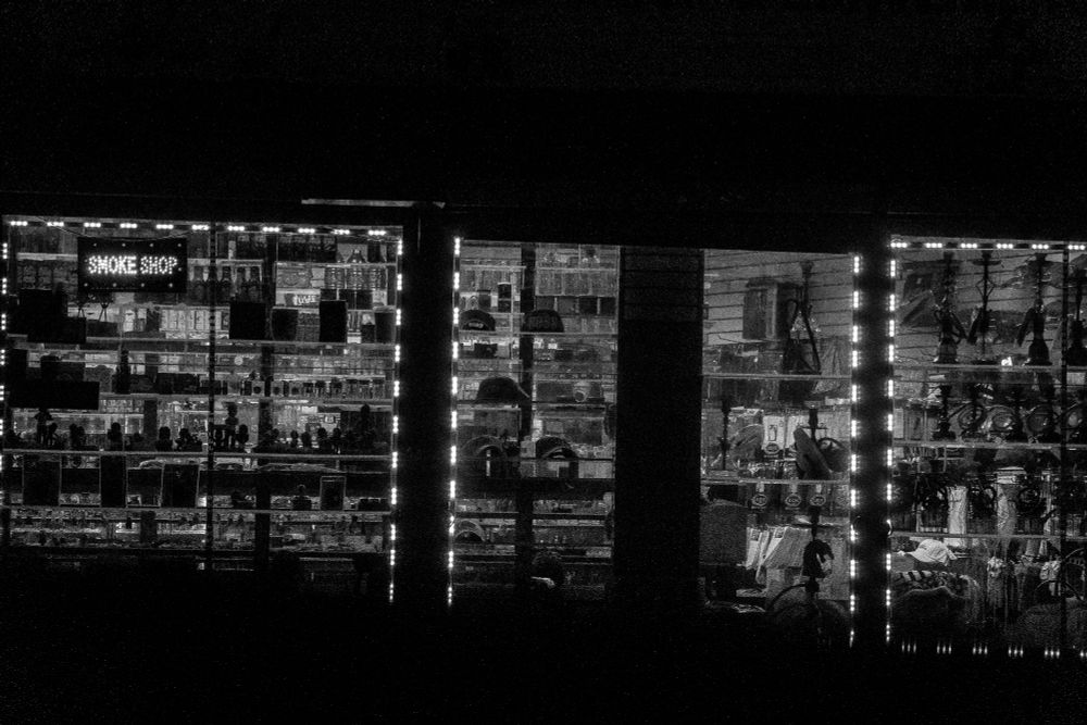 Interior of a brightly lit smoke shop at night. Shelves line the walls, stocked with pipes, hookahs, vapes, and smoking accessories. A glowing sign labeled “SMOKE SHOP” appears on the left. The space is neat and fully stocked, with products arranged in rows and clusters.  
