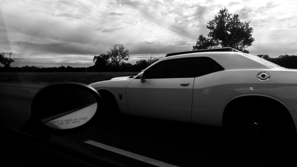 Black-and-white photo taken from inside a moving vehicle, showing the side mirror in the foreground with a warning text: “OBJECTS IN MIRROR ARE CLOSER THAN THEY APPEAR.” A white car drives parallel on the road, with overcast sky, trees, and power lines in the blurred background. The image captures motion, contrast, and perspective.  
