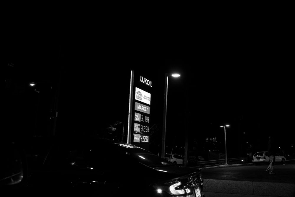 Nighttime view of a Lukoil gas station with prices lit up on a tall digital sign: Regular Cash – $3.19, Regular Credit – $3.29, Diesel – $4.59. A car is partially visible in the foreground, and a solitary pedestrian walks near the background. Streetlights cast a yellowish glow, while shadows and darkness edge the scene. The mood is stark, economical, and distinctly unromantic.
