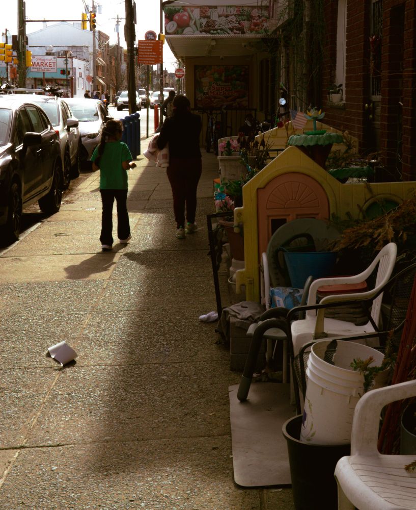 A woman and child walk down a sunlit sidewalk past chairs, buckets, and a playhouse. Their steps echo quiet defiance, everyday memory, and the rhythm of inherited language.
