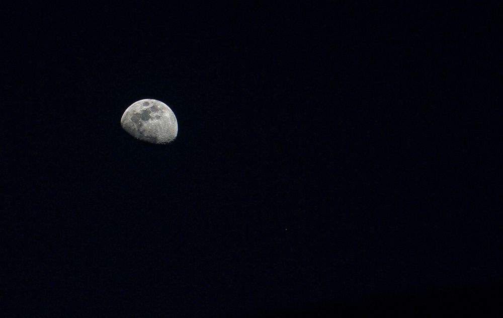 A close up of the moon in a dark night sky, captured in its waxing gibbous phase. The surface shows visible craters and textured shadows. One small star appears near the lower right, emphasizing the moon’s brightness and isolation. The scene feels quiet, precise, and suspended in orbital rhythm.
