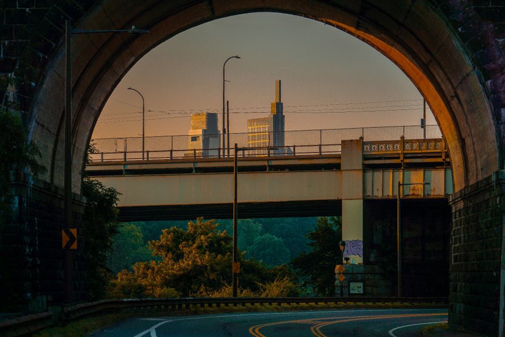 Morning sun casts dense orange hues across an urban bridge, held between stone archway and distant buildings. The orange is not ambient—it marks directional light, concentrated along railings and streetlamps. Vegetation below the bridge remains in shadow, holding contrast. The archway frames the scene as structural threshold, not ornament. The orange operates as temporal signal—early, exact, rising.

